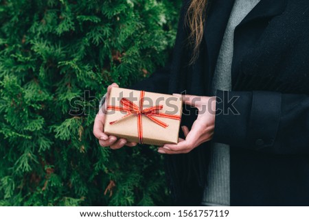 Woman giving a christmas or birthday gift wrapped in brown paper and red ribbon. Copy space. Thuja occidentalis background. Green Christmas texture.