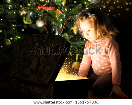 Child by Christmas tree opens magic chest. Portrait of little girl looking inside of gift box at home. Miracle and surprise for baby from Santa on New Year. Cute kid and present with fairy lights.