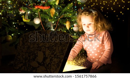 Kid by Christmas tree opens magic chest. Child looks inside of gift box at home. Miracle and surprise for baby from Santa on New Year. Cute little girl and present with fairy lights at winter night.
