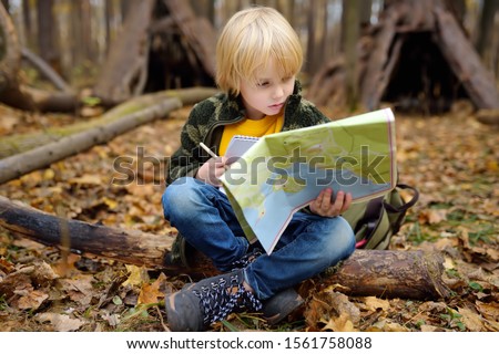 Little boy scout is orienteering in forest. Child is sitting on fallen tree and looking on map on background of teepee hut. Concepts of adventure, scouting and hiking tourism for kids.