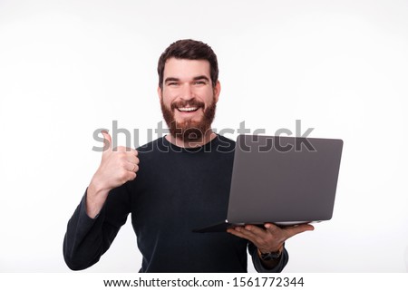 Photo of happy bearded guy holding a laptop while looking at camera and shoeing thumb uo, over white isolated background 