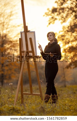 silhouette of beautiful young woman artist painting a picture on the easel, girl figure with a brush and a palette of colors on the background of autumn landscape, a concept of hobby and art
