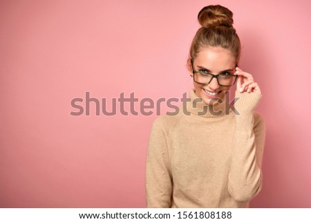 A girl with a high beam stands in a sweater on a pink background, smiles and looks to the side, holding glasses with her hand