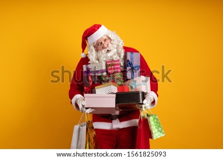 Male actor in a costume of Santa Claus holds a lot of gift boxes and packages in his hands and poses on a yellow background