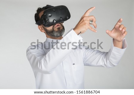 Handsome Arab man in VR headset enjoying experience and gesturing while standing against white background. Royalty-Free Stock Photo #1561886704