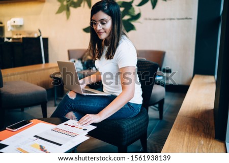Side view of young casual female freelance designer using color swatches while sitting at workplace and working with laptop on new web design 