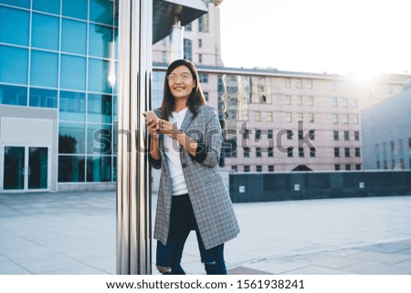 Happy woman in casual outfit and spectacles smiling and thinking while using smartphone near pillar and looking away on city street