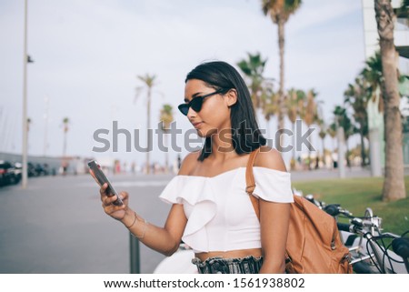 Side view of millennial blogger in fashionable sunglasses connecting to 4g wireless for tracking location GPS during time for walk on city street, attractive Latino woman installing application
