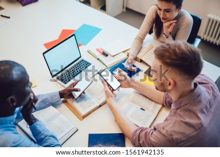 From above glad diverse coworkers in casual wear brainstorming while sitting at white table in light modern office and browsing laptop with smartphone on blurred background