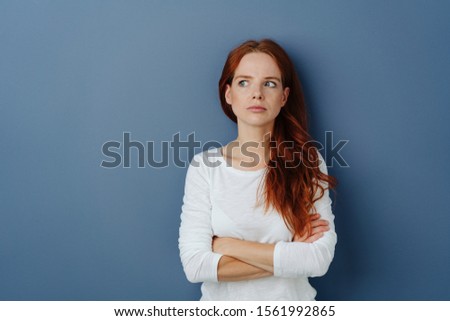Thoughtful jealous young woman looking aside with folded arms and a speculative expression over a blue studio background with copy space Royalty-Free Stock Photo #1561992865