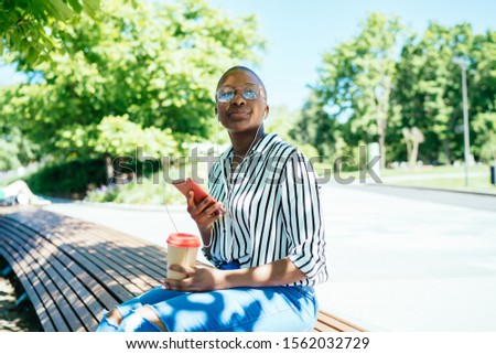 Gorgeous black female sitting outside in bright sunlight holding cup drinking coffee and listening music from mobile device looking away focused