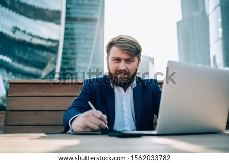 Executive concentrated bearded guy in formal wear writing ideas for remote work in notebook watching laptop sitting at table in park of modern city