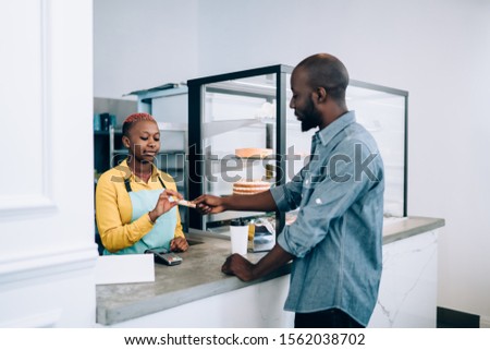 Pensive African American customer giving credit card to paying for beverage to go and food to attentive seller while standing beside counter in modern bakery shop Royalty-Free Stock Photo #1562038702