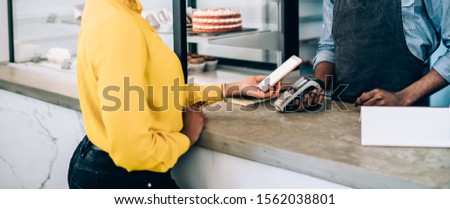 Low angle happy African American seller holding terminal and waiting while client making contactless payment with smartphone in light modern pastry shop
