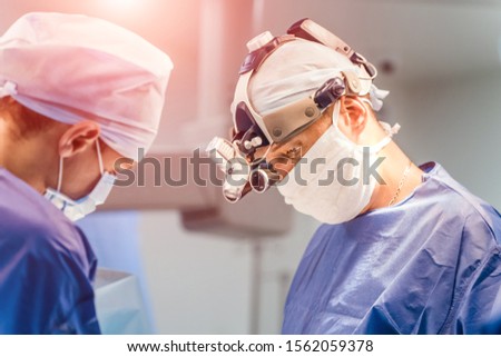 Specialists in medical uniform are doing an operation in hospital. Surgeon with magnifying glass on his head is working with assistants in the operating room.
