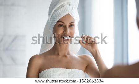 Happy healthy young woman brushing white teeth look in mirror, smiling beautiful lady with towel on head hold toothbrush clean mouth with toothpaste in bathroom, morning hygiene dental care concept