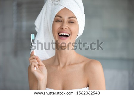 Cheerful young woman with towel on head holding toothbrush laughing look at camera, attractive happy lady with healthy white teeth dental smile do cleaning tooth morning routine in bathroom, portrait