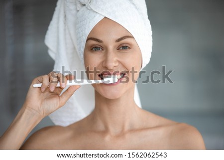 Happy young woman with white towel on head brushing healthy teeth look at camera, smiling attractive lady hold toothbrush clean mouth with toothpaste in bathroom, morning dental care concept portrait