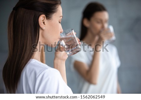 Thirsty young woman hold glass drink fresh clean water reflecting in mirror, dehydrated healthy lady hydrate thirst in morning keep healthcare balance for skin body care concept, over shoulder view