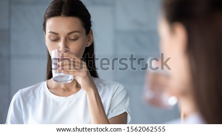 Attractive young healthy woman hold glass drink fresh mineral water in morning, thirsty lady feel thirst keep hydration diet nutrition weightloss balance concept stand in bathroom reflect in mirror