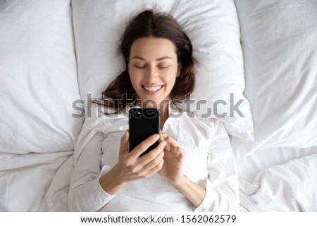 Happy young woman hold smart phone lying awake in bed in morning, smiling lazy attractive lady using social media apps check messages texting on smartphone relax in bedroom with cellphone, top view