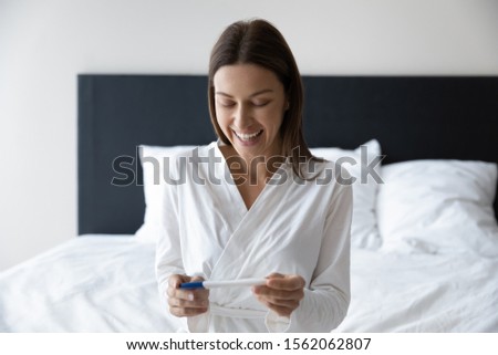 Happy beautiful pregnant young woman wear night gown hold positive result pregnancy test sit on bed in morning, excited surprised lady expect baby after infertility treatment in vitro fertilization