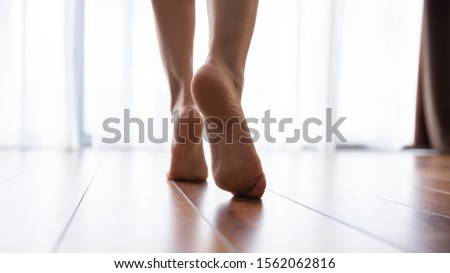 Female feet walking on warm heated floor close up view, barefoot girl legs going on hardwood living room wooden flooring at modern home house, domestic underfloor heating concept, close up rear view Royalty-Free Stock Photo #1562062816