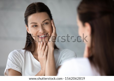 Happy beautiful young lady touching face look in bathroom mirror, happy pretty woman enjoy healthy soft moisturized hydrated skin, facial skincare rejuvenation moisture spa everyday treatment concept Royalty-Free Stock Photo #1562062819