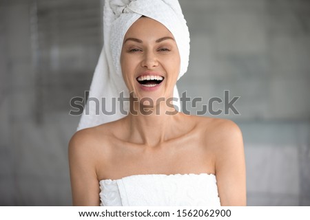 Positive young lady wrapped in towels laughing looking at camera, happy beautiful 30s woman with white teeth dental smile healthy face skin posing in bathroom after sauna morning routine, portrait