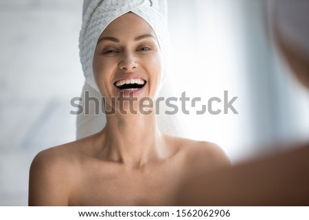 Happy young woman with towel on head laughing looking in mirror camera, cheerful attractive happy lady with white teeth dental smile healthy skin having fun do morning routine in bathroom, portrait