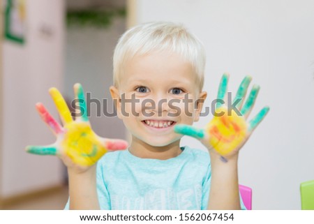 Close up of a cheerful little boy showing painted hands at the camera while playing in the kindergarten