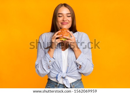 Junk Food. Cheerful Woman Smelling Burger Standing Over Yellow Background. Studio Shot