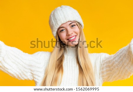 Selfie concept. Pretty young woman in knitted white hat holding camera, taking selfie and smiling, standing against yellow background