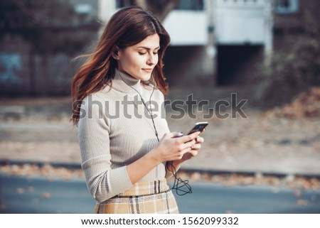 Image of beautiful stylish woman holding mobile phone. Young european girl standing at the street and using cellphone. Woman listening to music with the phone and having fun. Mobile internet concept. 