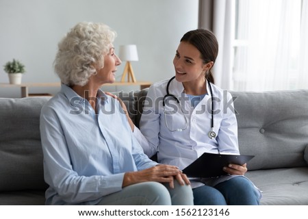 Caring young woman nurse help old granny during homecare medical visit, female caretaker doctor talk with elder lady give empathy support encourage patient sit on sofa older people healthcare concept Royalty-Free Stock Photo #1562123146