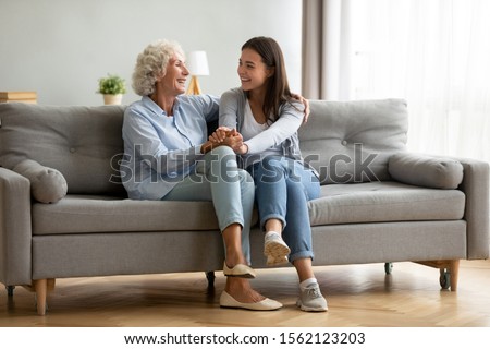 Happy adult granddaughter and senior grandmother having fun enjoying talk sit on sofa in modern living room, smiling old mother hugging young grown daughter bonding chatting relaxing at home together Royalty-Free Stock Photo #1562123203