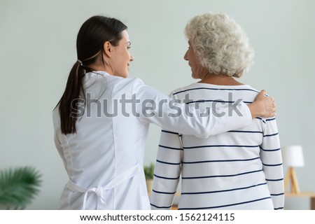 Rear back view of young female doctor physiotherapist caretaker help injured old adult grandma patient assisting holding walking with senior lady at home hospital, elder people physiotherapy concept Royalty-Free Stock Photo #1562124115