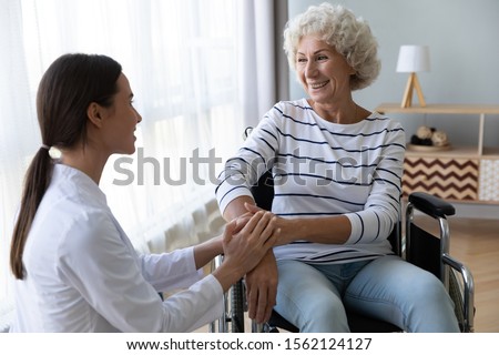 Caring female nurse caregiver hold hand support disabled elder woman patient sit on wheelchair at home hospital, young doctor carer help paralyzed old granny on wheel chair, senior homecare concept Royalty-Free Stock Photo #1562124127