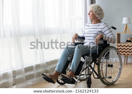 Pensive disabled senior grandma patient sit on wheelchair alone at home hospital look through window, thoughtful sad old woman feel depressed lonely, retired people disability and loneliness concept Royalty-Free Stock Photo #1562124133
