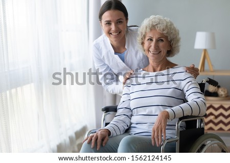 Happy disabled handicapped grandma patient sit on wheelchair and young nurse caregiver look at camera at home hospital, female paramedic helping senior paralyzed woman, eldercare concept, portrait
