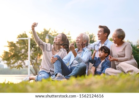 three generation happy asian family sitting on grass taking a selfie using mobile phone outdoors in park Royalty-Free Stock Photo #1562124616
