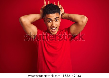 Young brazilian man wearing t-shirt standing over isolated red background Posing funny and crazy with fingers on head as bunny ears, smiling cheerful