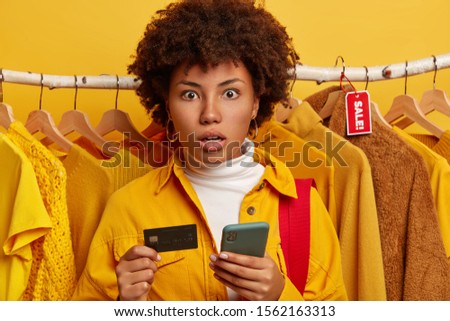 Amazed lady with Afro hairstyle, dressed in yellow shirt, poses over clothing racks, holds modern cellular and credit card, yellow background. People, shopping, consumerism, technology concept
