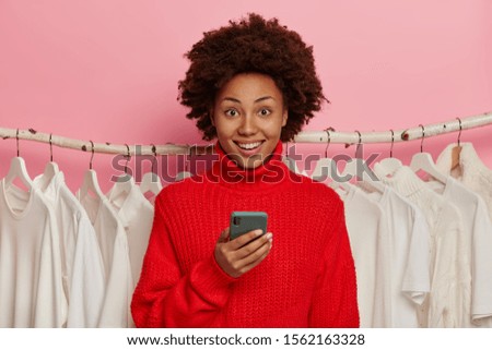 Happy dark skinned woman dresses red sweater, stands against new collection of clothes, uses mobile phone for surfing internet, smiles broadly, isolated over pink wall. Many white outfits on hangers