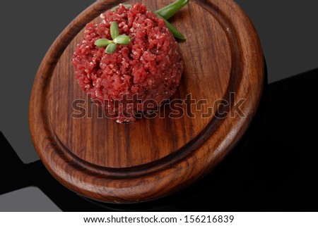 very big raw hamburger cutlet with sprouts and chili pepper on wooden plate over black background