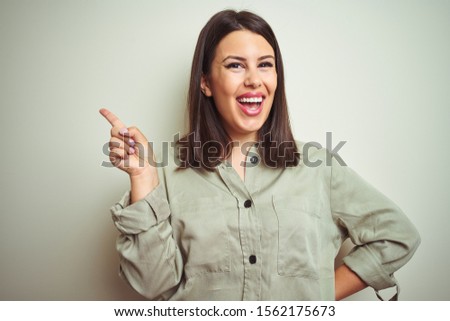 Young beautiful brunette woman wearing green shirt over isolated background with a big smile on face, pointing with hand and finger to the side looking at the camera.