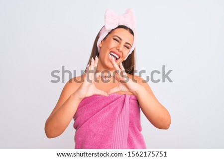 Beautiful young woman wearing pink shower towel and beauty headband over white background smiling in love doing heart symbol shape with hands. Romantic concept.