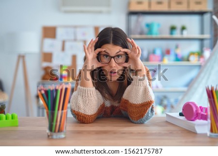 Young beautiful teacher woman wearing sweater and glasses sitting on desk at kindergarten Trying to open eyes with fingers, sleepy and tired for morning fatigue
