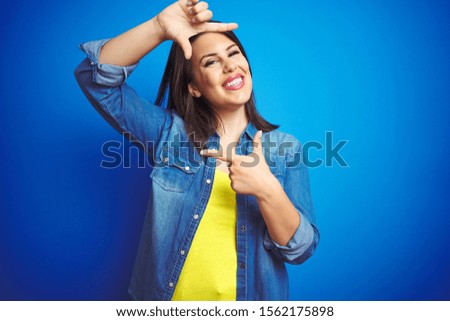 Young beautiful brunette woman wearing casual blue denim jacket over blue isolated background smiling making frame with hands and fingers with happy face. Creativity and photography concept.