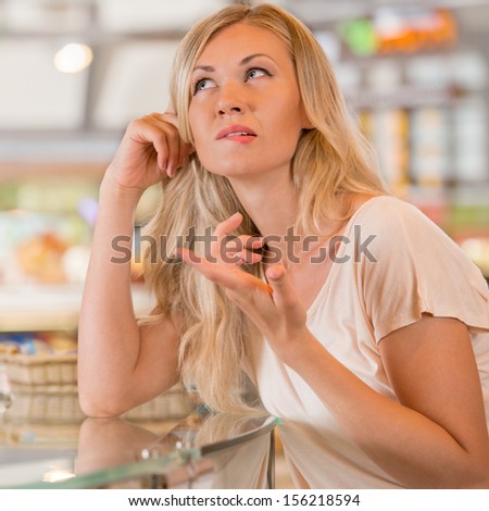 Beautiful young woman standing near supermarket showcase full of different products and deciding what to buy